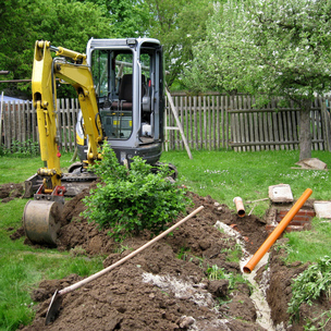 Kleiner gelber Bagger steht in einem Garten mit aufgebaggerter Wiese, um Rohre verlegen zu können.